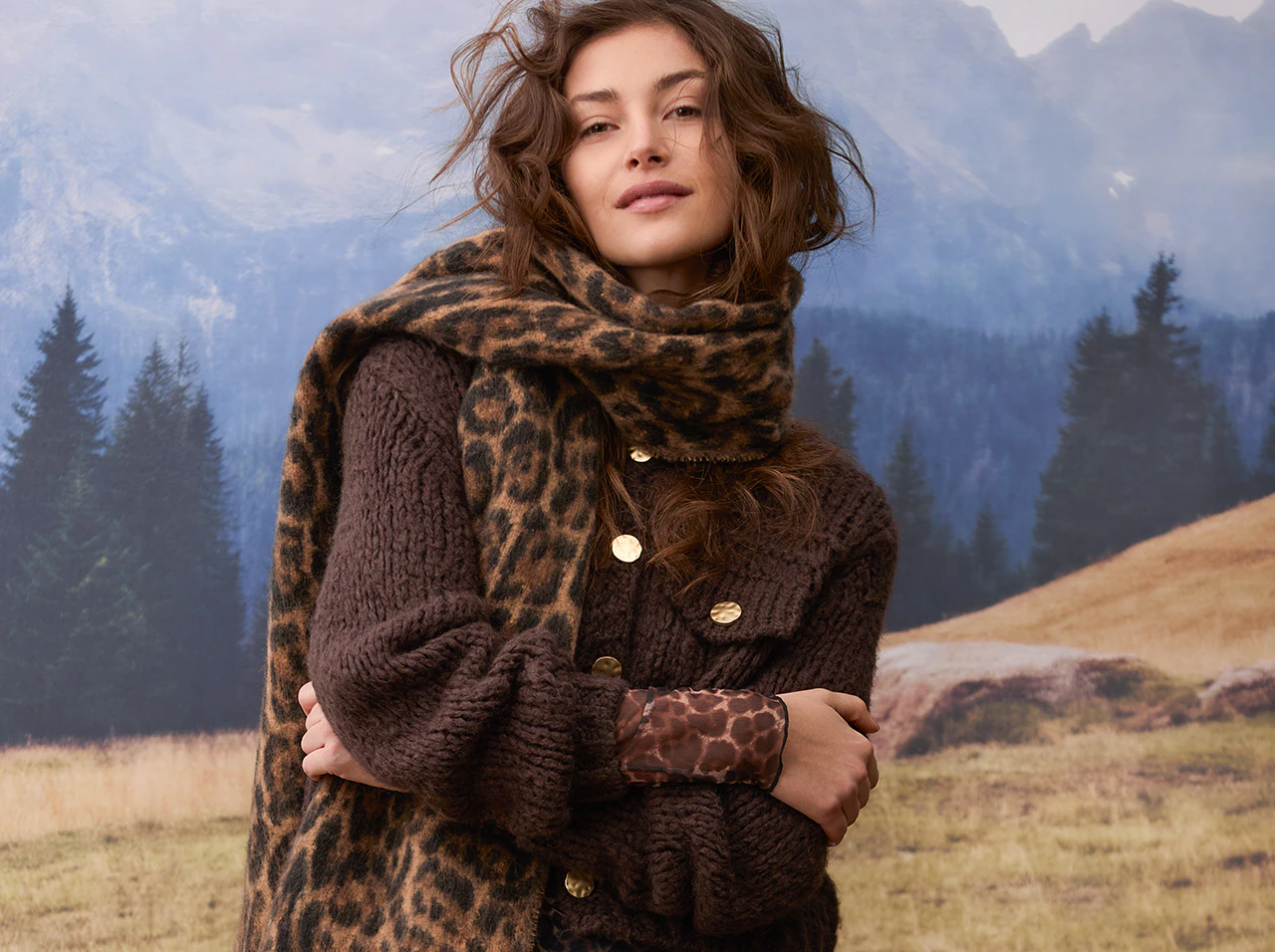 Model trägt eine dunkelbraune Strickjacke mit goldfarbenen Knöpfen, einen Leo-Schal und ein gemustertes Oberteil.