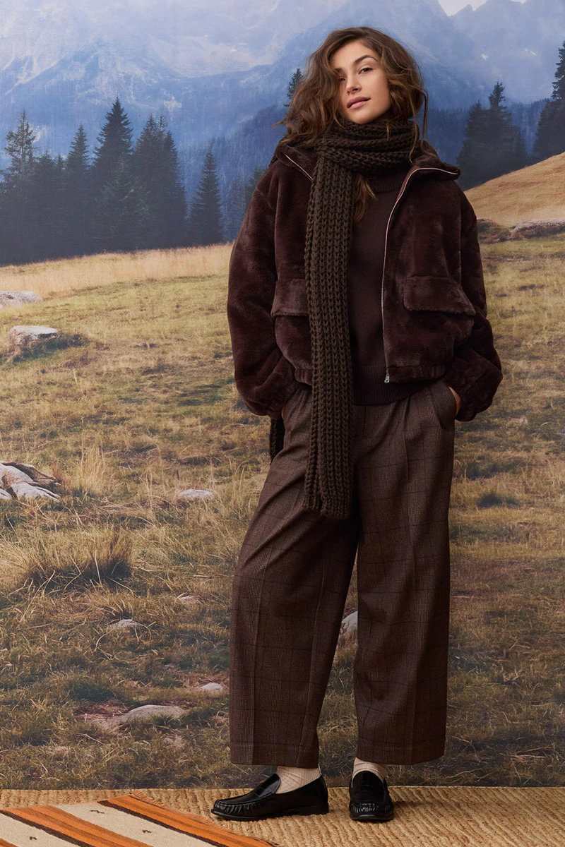 Autumn women’s fashion: A model is wearing a dark brown jacket with a large knitted scarf, a brown jumper and loose-fitting checked trousers. She is wearing black loafers. In the background, an alpine mountain landscape with woods, meadows and rocks can be seen.