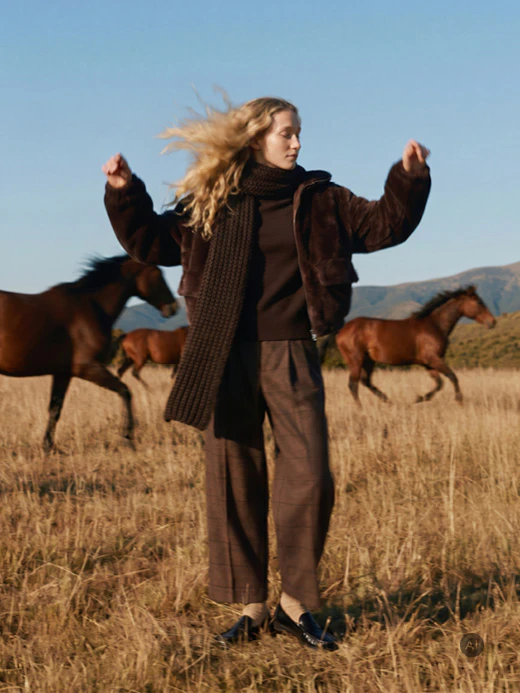 Herbstliche Damenmode: Model trägt eine braune Jacke, einen dunklen Strickpullover mit langem Schal, eine braune karierte Hose und schwarze Loafer. Sie steht auf einem Feld, während im Hintergrund mehrere Pferde laufen und Berge unter blauem Himmel zu sehen sind.
