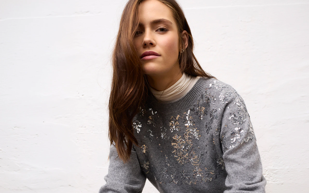 A woman wearing a grey jumper with sparkling sequinned details over a cream-coloured turtleneck top.