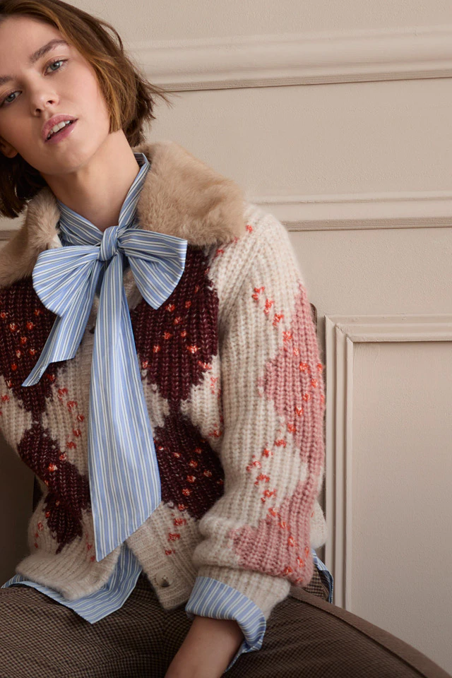 The model is wearing a patterned knitted jumper over a striped blouse with a bow and a checked skirt. A brown dog is sitting next to her
