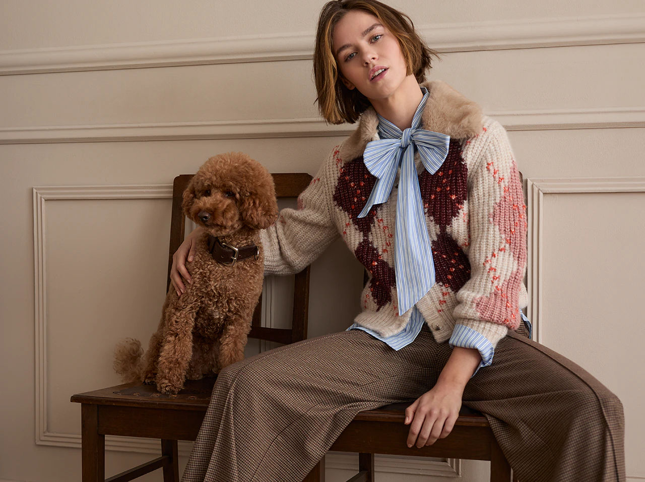 Model trägt einen gemusterten Strickpullover über einer gestreiften Bluse mit Schleife und einen karierten Rock. Neben ihr sitzt ein brauner Hund.