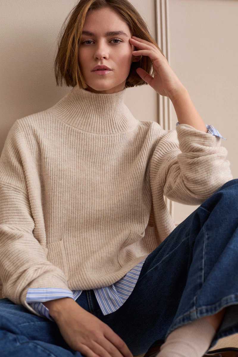 The model is wearing a cream-coloured knitted jumper with a stand-up collar over a blue and white striped blouse and dark jeans.