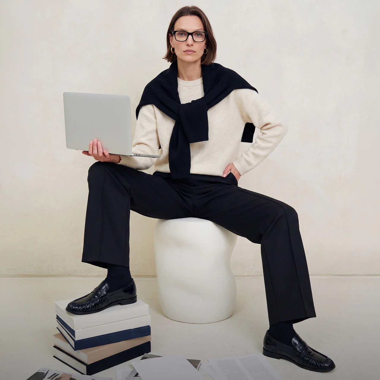 A model wearing a cream-coloured jumper with a dark blue jumper draped over her shoulders, black trousers and black loafers with ribbed socks.