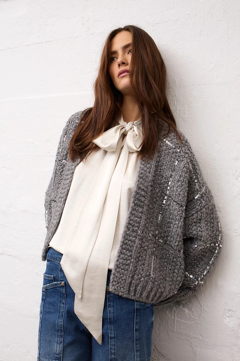 A grey women’s cardigan with sequins, a cream-coloured bow-neck blouse and blue jeans from Oui.
