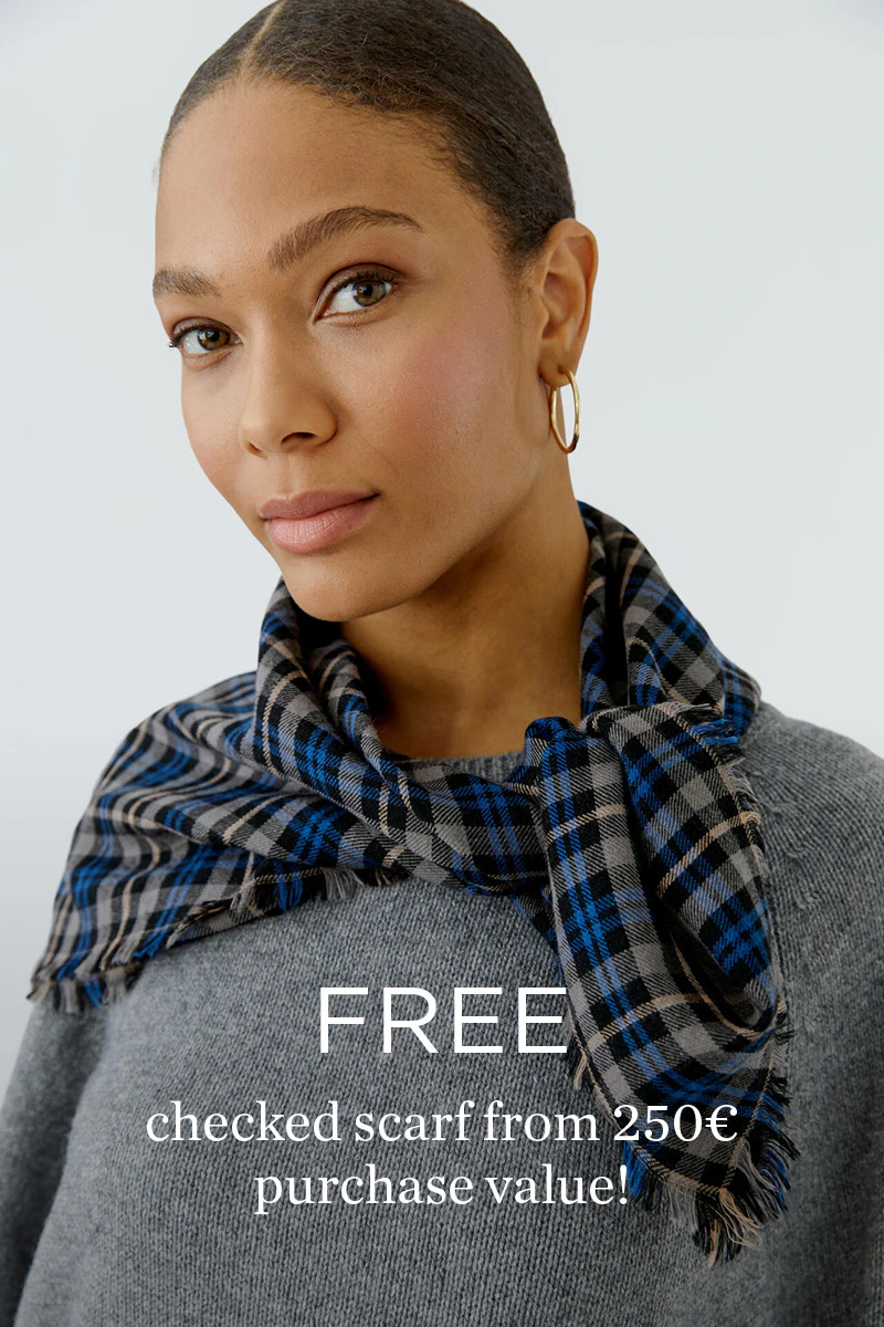 Model wearing a grey knitted jumper with a blue-and-grey checked scarf from the Oui Heritage Reset collection - accompanied by the message "Get a free checked scarf with a purchase of €250 or more".