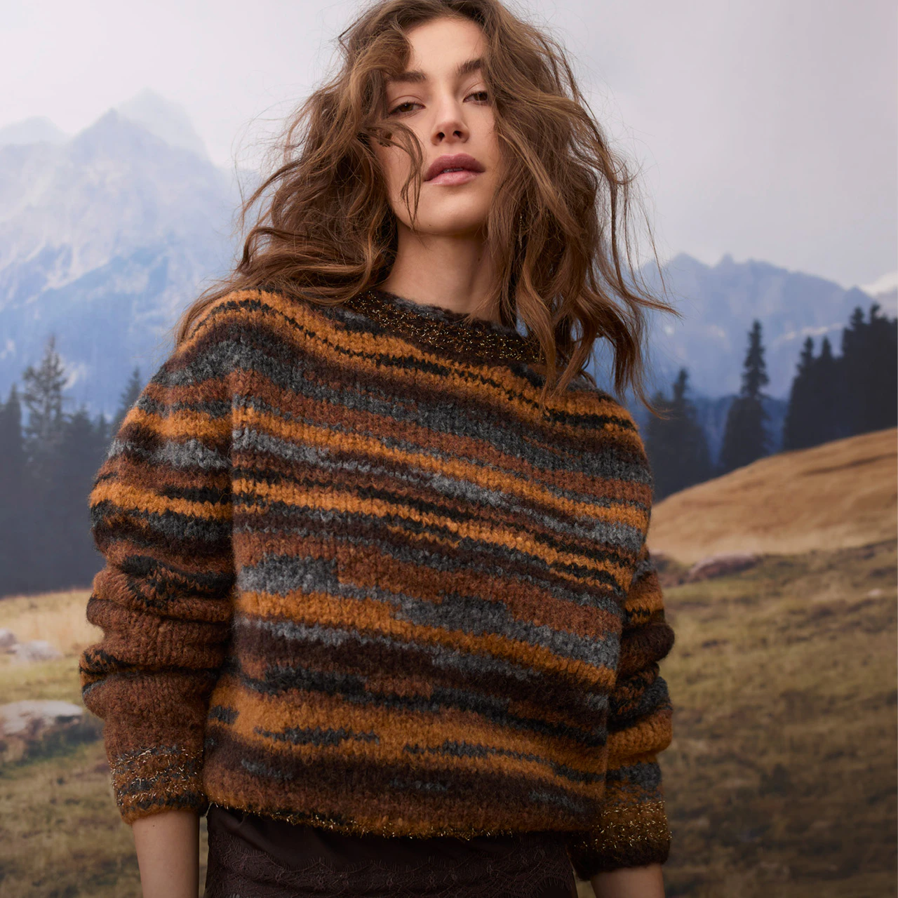 The model is wearing a striped knitted jumper in warm shades of brown, orange and grey, with subtle glittery details.