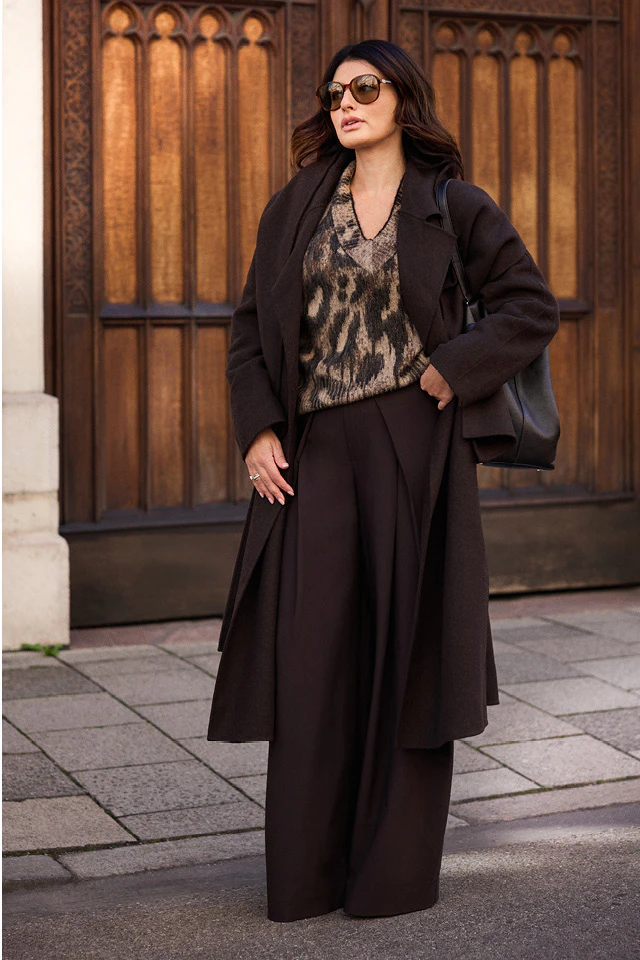 Influencerin Sofiya Balci trägt einen dunkelbraunen Mantel und eine weite Hose im Ton-in-Ton-Look, kombiniert mit einem gemusterten Pullover im Animalprint, Sonnenbrille und schwarzer Schultertasche. Der urbane Look wird vor einer kunstvoll verzierten Holztür in Szene gesetzt.