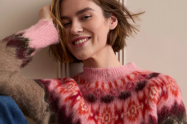 The model is wearing a brown Norwegian-style knitted jumper with a pink-and-red pattern and blue jeans.