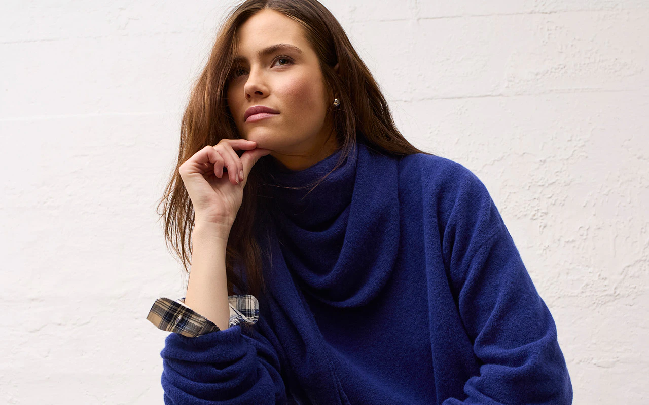 The model is wearing a blue oversized jumper with checked cuffs and a checked shirt underneath.