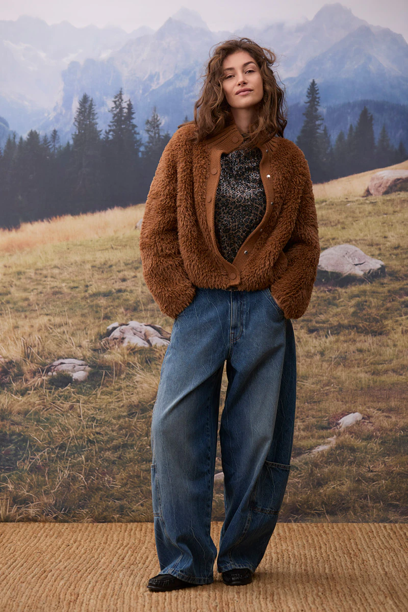 Autumn women’s fashion: a model is wearing a brown teddy jacket over a dark patterned jumper, paired with loose-fitting blue jeans and black shoes. In the background, an alpine mountain landscape can be seen, featuring woods, meadows and rocks.