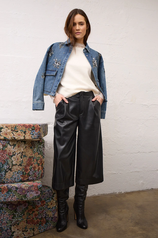 A woman wearing a denim jacket draped over her shoulders with glittery details, a beige jumper and black 7/8-length trousers with a leather-effect finish.