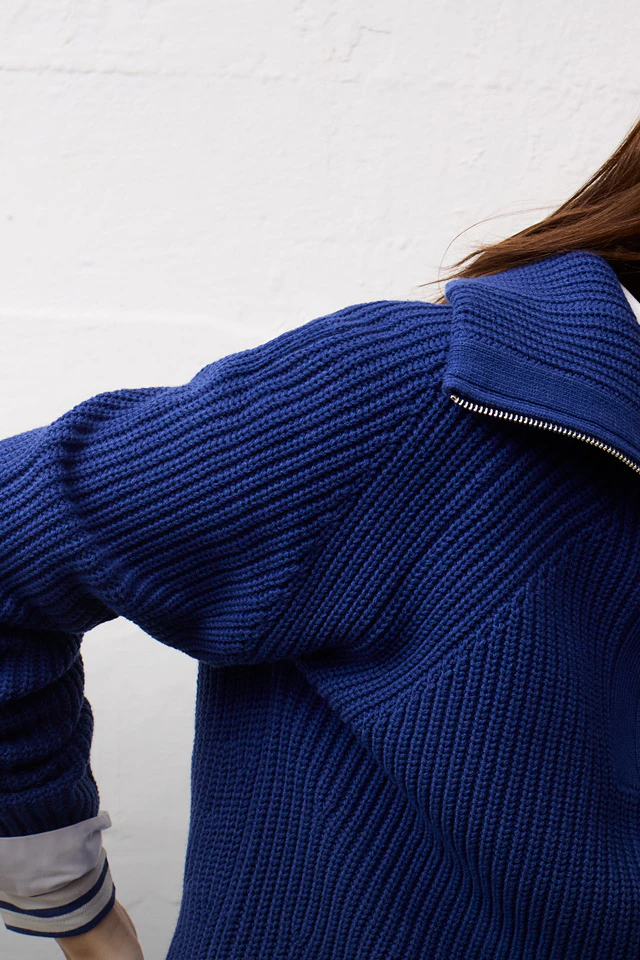 A woman in a blue knitted jumper with a zip and a white shirt with striped cuffs, leaning casually against a retro television.