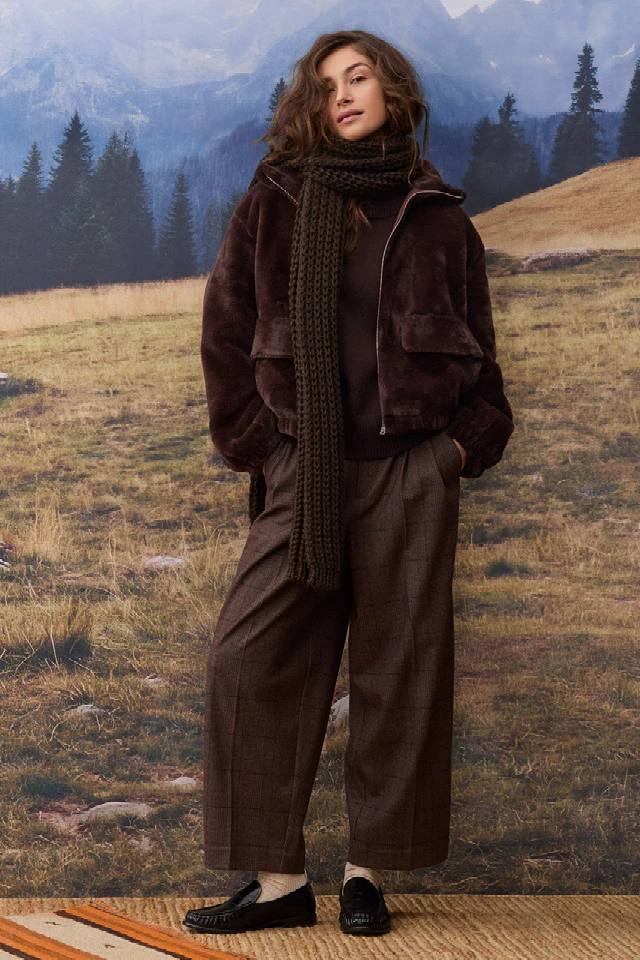 The model is wearing a dark brown jacket with a large knitted scarf, a brown jumper and loose-fitting checked trousers.