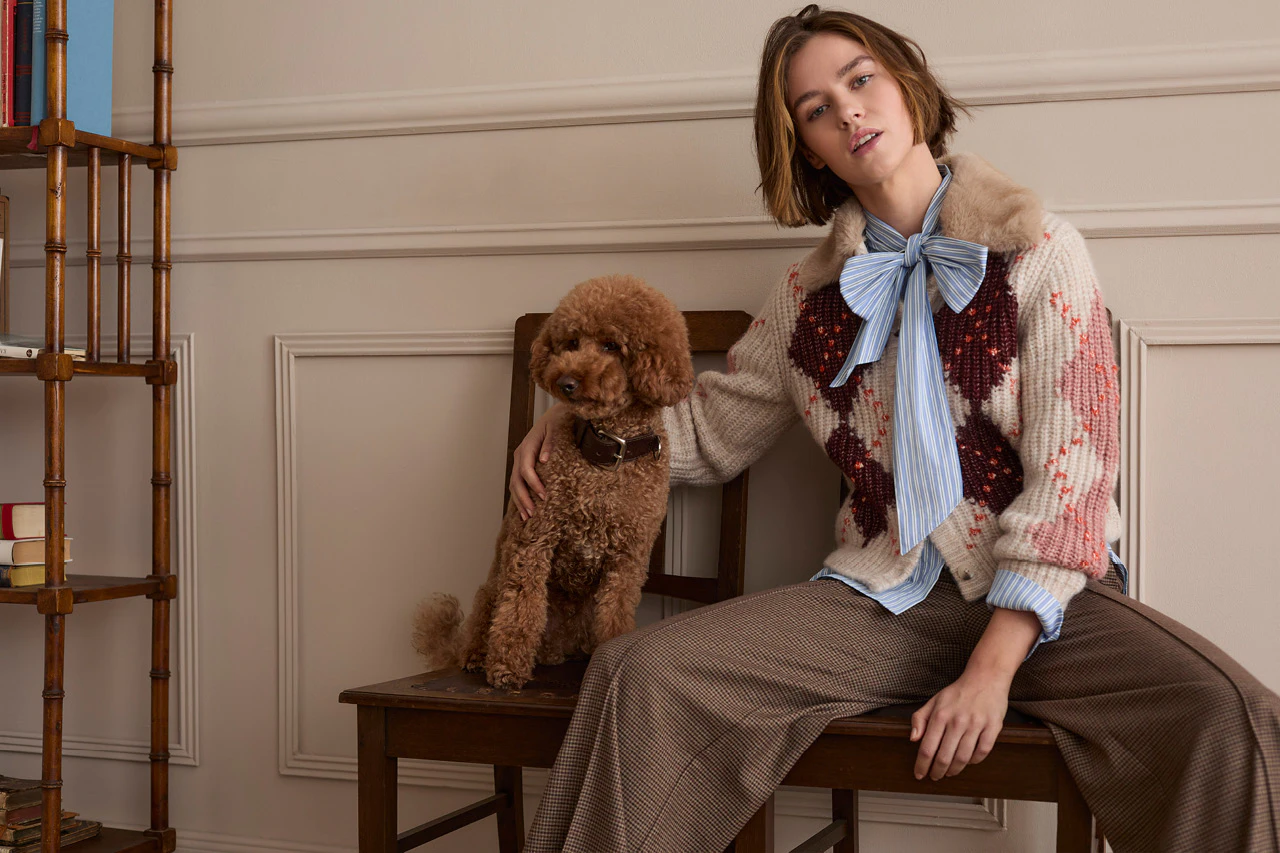 The model is wearing a patterned knitted jumper over a striped blouse with a bow and a checked skirt. A brown dog is sitting next to her.