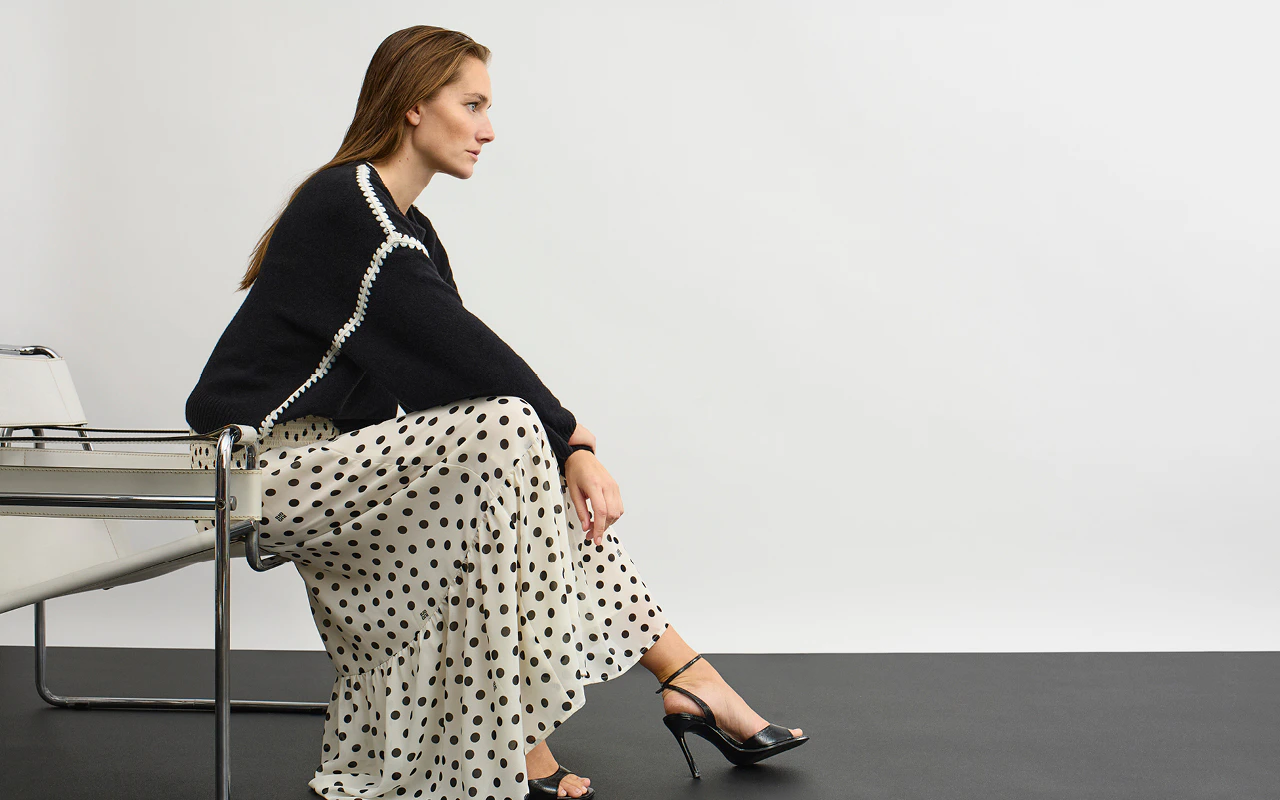 A woman wearing a black jumper with contrasting white stitching and a white, loose-fitting midi skirt with black dots.