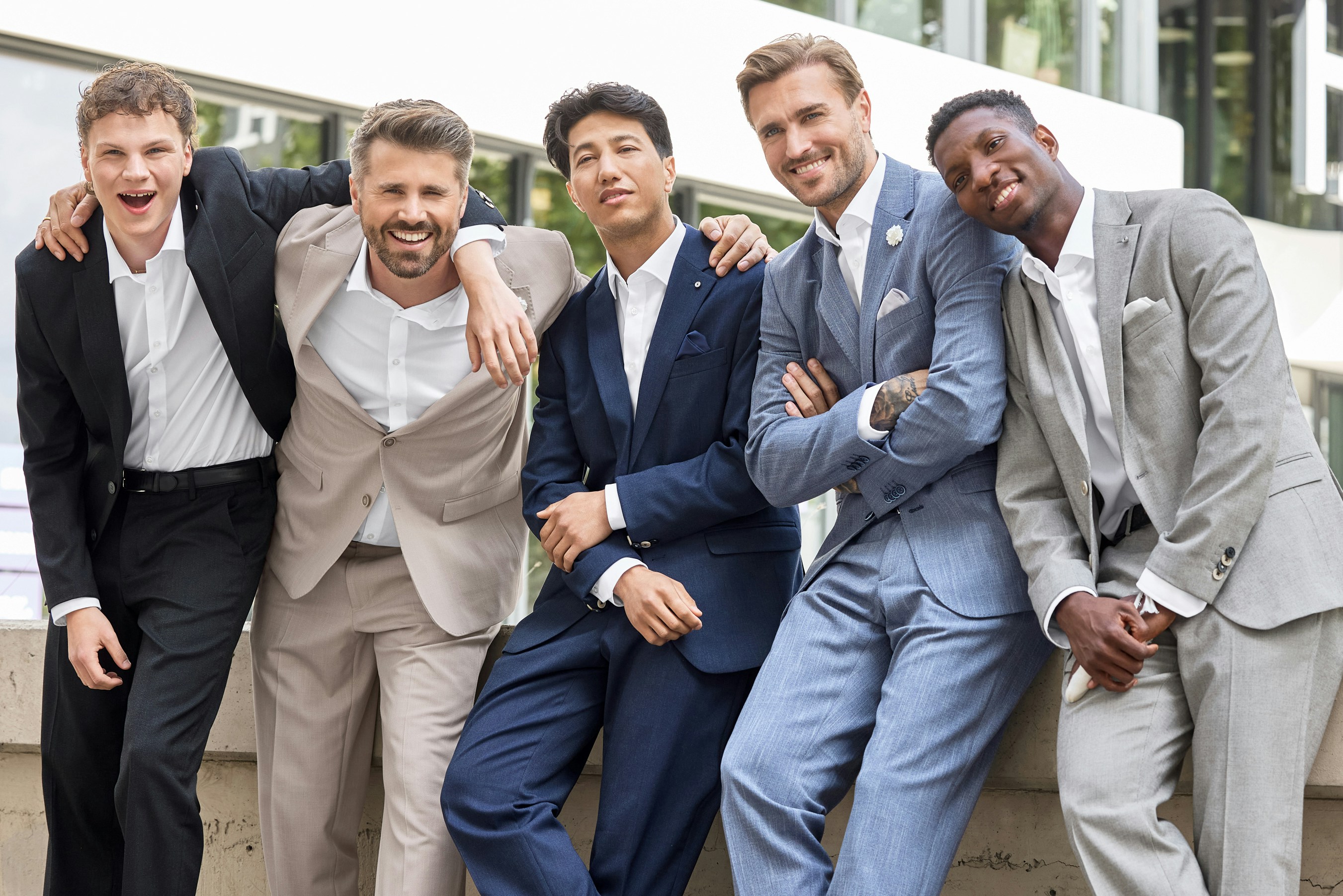 Fünf Männer tragen Anzüge in Schwarz, Beige, Blau und Grau und lehnen nebeneinander an einer Mauer im Außenbereich