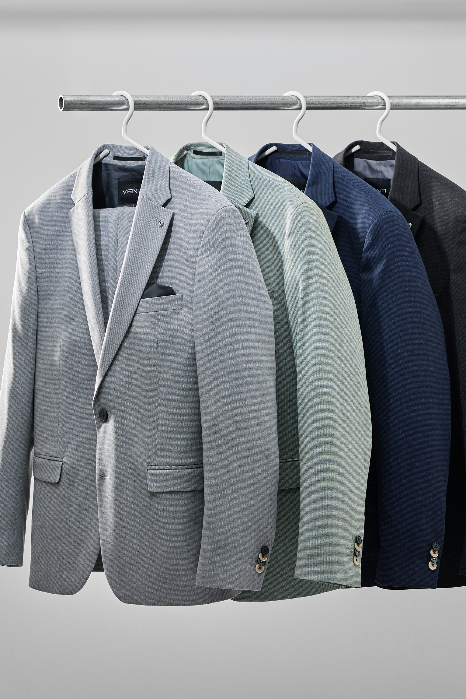Mehrere Herrenanzug-Sakkos in Grau-, Grün- und Blautönen hängen nebeneinander auf einer Kleiderstange
