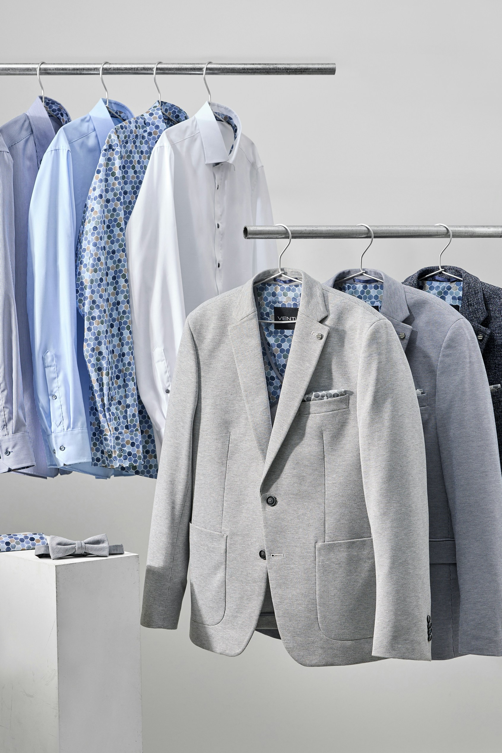 Weiße und blaue Hemden sowie graue Herrenanzug-Sakkos hängen auf zwei Kleiderstangen, gemusterte Innenstoffe sind sichtbar
