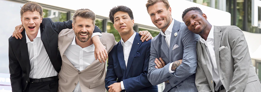 Fünf Männer tragen Anzüge in Schwarz, Beige, Blau und Grau und lehnen nebeneinander an einer Mauer im Außenbereich