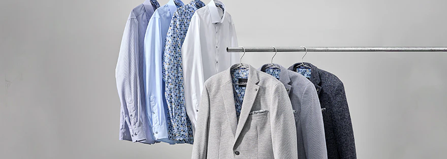 Weiße und blaue Hemden sowie graue Herrenanzug-Sakkos hängen auf zwei Kleiderstangen, gemusterte Innenstoffe sind sichtbar