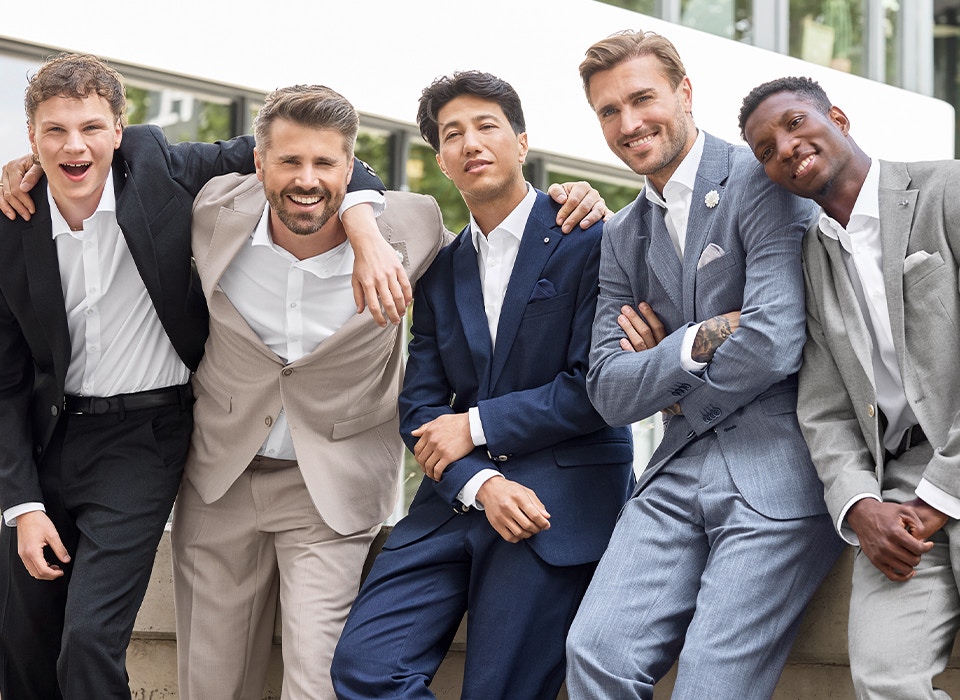 Fünf Männer tragen Anzüge in Schwarz, Beige, Blau und Grau und lehnen nebeneinander an einer Mauer im Außenbereich