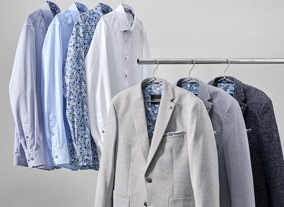 Weiße und blaue Hemden sowie graue Herrenanzug-Sakkos hängen auf zwei Kleiderstangen, gemusterte Innenstoffe sind sichtbar
