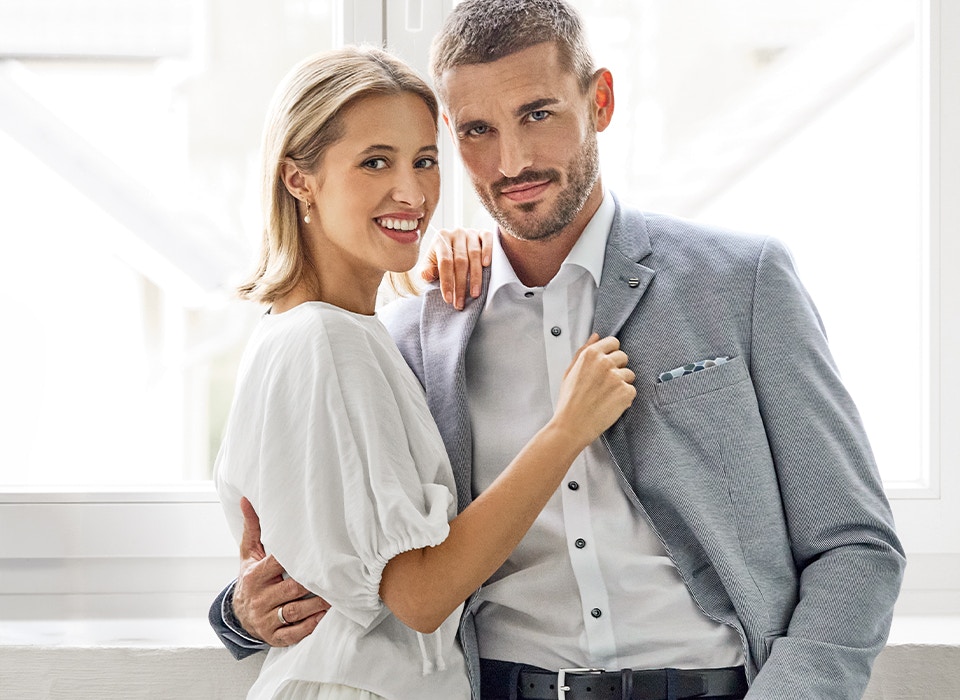 Mann in hellem Sakko mit weißem Hemd steht neben Frau in weißem Kleid, beide blicken in die Kamera