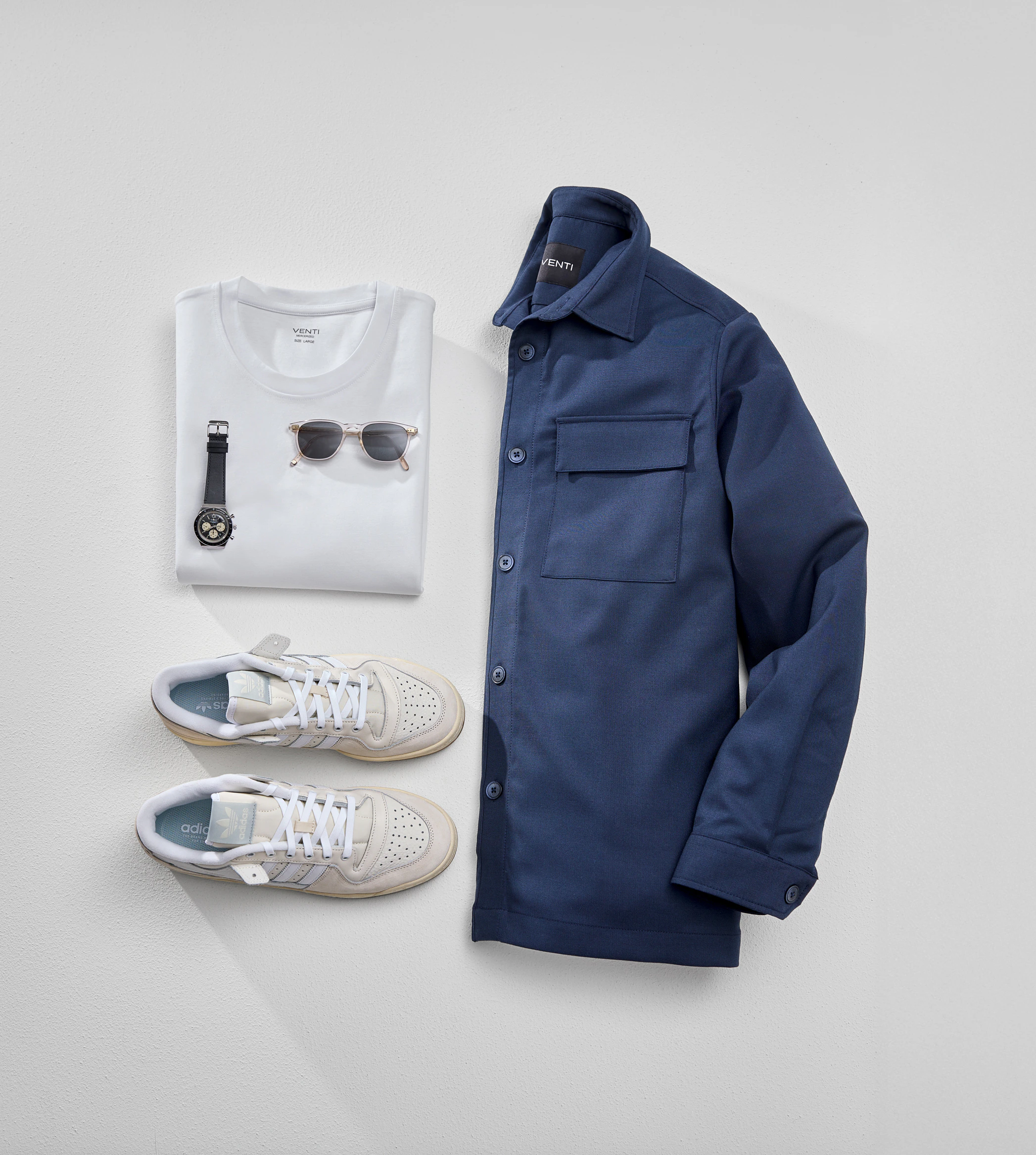 Blaue Hemdjacke liegt neben einem weißen T-Shirt und beigen Sneakern. Auf dem T-Shirt liegt eine Sonnenbrille und eine Uhr.
