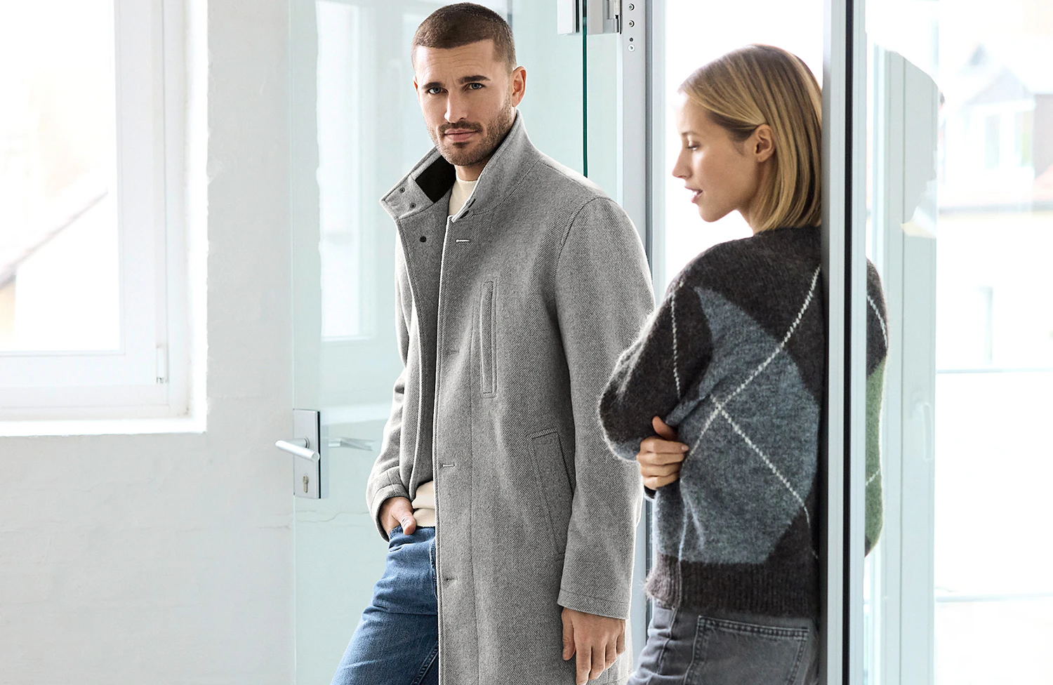 Mann in einem eleganten grauen Mantel und blauer Jeans neben einer Frau im gemusterten Strickpullover und grauer Jeans in einem hellen, modernen Ambiente.