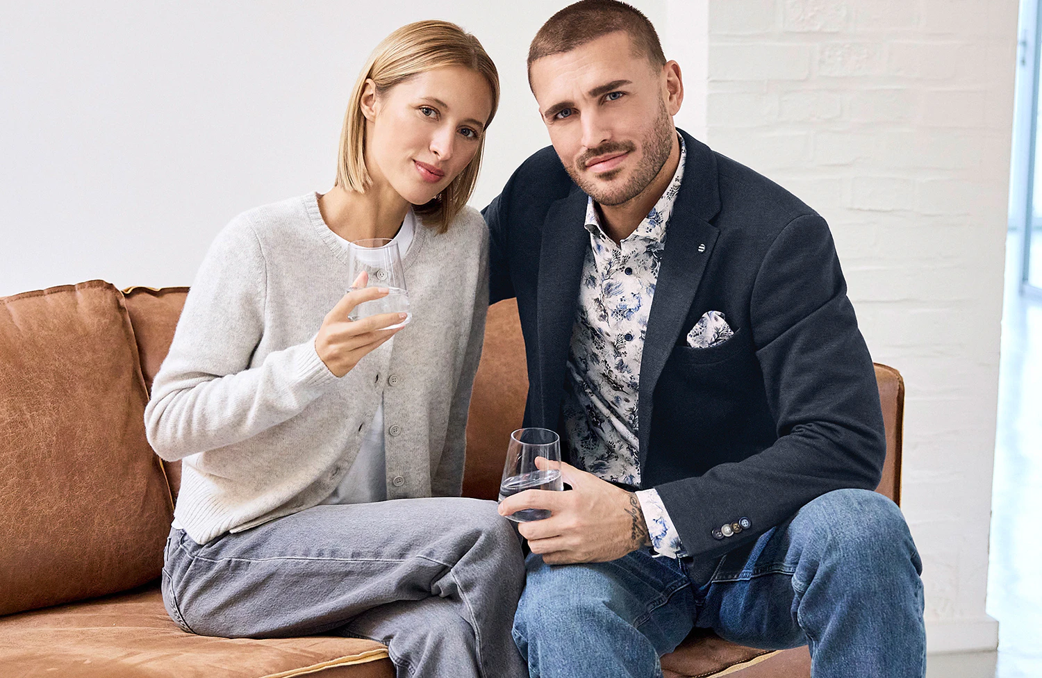 Mann im dunkelblauen Sakko mit floral gemustertem Hemd und blauer Jeans sitzt neben einer Frau in hellgrauer Strickjacke auf einem braunen Ledersofa.